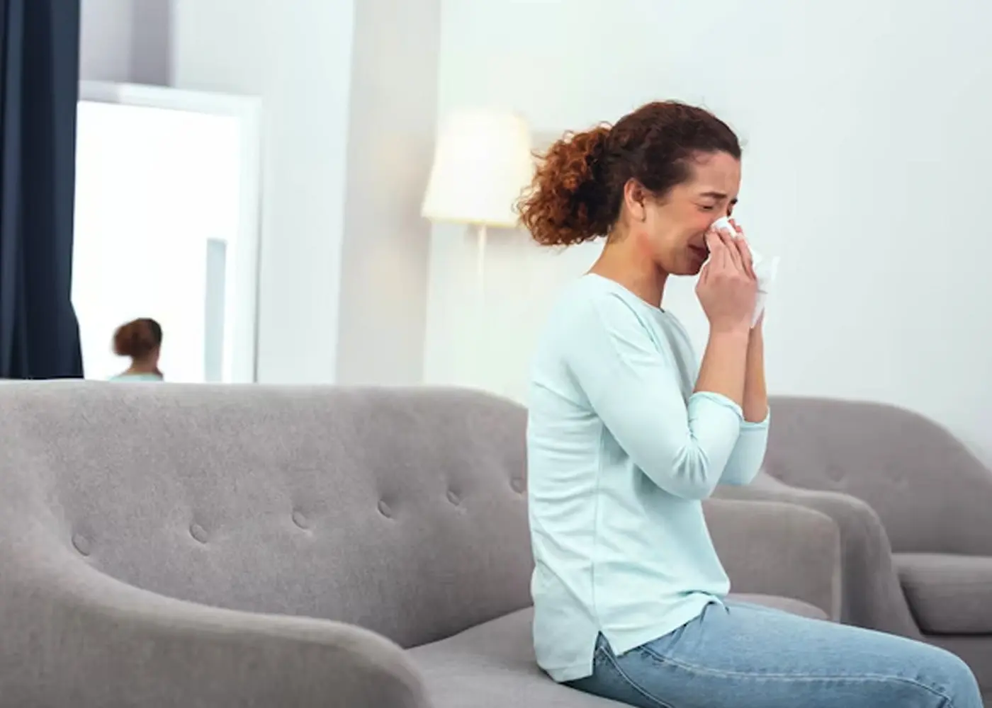 A woman sitting on a couch and sneezing, representing the need for hay fever treatment.